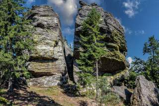 Zwei große, verwitterte Felsformationen stehen dicht nebeneinander, umgeben von grünem Nadel- und Laubwald unter blauem Himmel mit einigen Wolken.