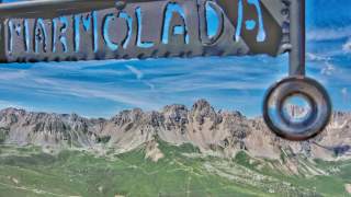 Blick auf die markanten, felsigen Gipfel der Marmolata mit grünen Almwiesen im Vordergrund und einem blauen Himmel darüber, im Vordergrund ein Metallrahmen mit der Aufschrift „MARMOLADA“.