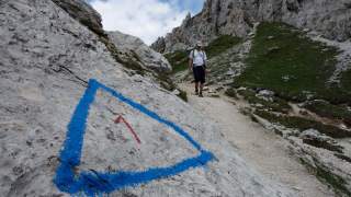 Wanderer auf einem Bergweg, im Vordergrund ein Felsen mit blauem Dreieck und roter Zahl 1 als Wegmarkierung. Im Hintergrund steile Felswände und bewölkter Himmel.