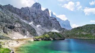 Klarer Bergsee mit grün-blauem Wasser vor felsigen, teils schneebedeckten Gebirgswänden, unter blauem Himmel mit vereinzelten Wolken, links am Ufer kleine Gruppen von Menschen.