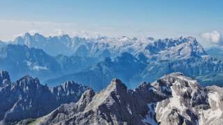 Blick auf eine Berglandschaft mit schroffen, teils schneebedeckten Gipfeln unter klarem blauem Himmel. Im Hintergrund sind mehrere Bergketten mit leichten Wolken sichtbar.