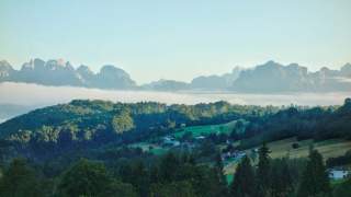 Landschaft mit grünen Wäldern und Wiesen vor einer Bergkette, darüber eine Schicht Nebel oder Wolken. Vereinzelt stehen Häuser auf den Hügeln.