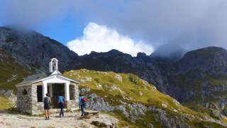 Drei Wanderer stehen vor einer kleinen Kapelle inmitten einer bergigen Landschaft mit teils wolkenverhangenem Himmel.