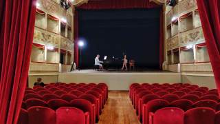Blick von den roten Theatersesseln auf eine Bühne, auf der ein Mann Klavier spielt und eine Frau daneben steht, umgeben von einem historischen Zuschauerraum mit Logen und roten Vorhängen.