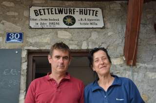 Zwei Personen stehen vor dem Eingang der Bettelwürf-Hütte, deren Schild über der Tür befestigt ist.