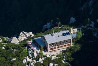 Berghütte mit Terrasse und Sonnenschirmen, umgeben von Felsen und Wald.