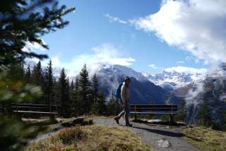 Wanderer mit Rucksack und Hut auf einem Bergpfad mit zwei Bänken, umgeben von Nadelbäumen, vor schneebedeckten Bergen und blauem Himmel.