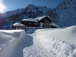 Verschneiter Weg führt zu einem Chalet-ähnlichen Haus in den Bergen, umgeben von hohen, verschneiten Hügeln und bewaldeten Bergen im Hintergrund bei Sonnenschein.