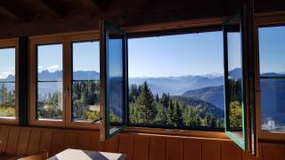 Blick aus einem geöffneten Fenster auf bewaldete Hügel und eine Bergkette unter blauem Himmel.