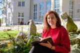 Frau mit rotbraunen Haaren und rotem Pullover sitzt draußen in der Sonne auf Steinen, hält ein Notizbuch und lächelt in die Kamera; im Hintergrund ein Gebäude mit großen Fenstern.