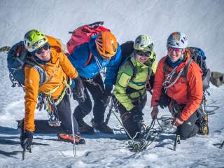 Vier Bergsteiger in bunter Ausrüstung und Helmen knien auf schneebedecktem Untergrund und sind mit Seilen gesichert.