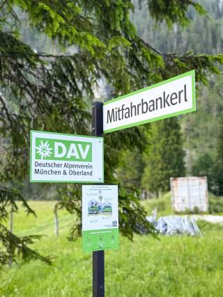 Schild mit der Aufschrift „Mitfahrbankerl“ und Hinweis auf den Deutschen Alpenverein, Sektion München & Oberland vor einem bewaldeten Gebiet.