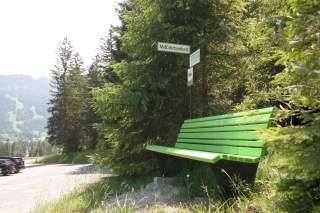 Eine grüne Bank an einem Wanderparkplatz in den Bergen. Daneben ein Schild 