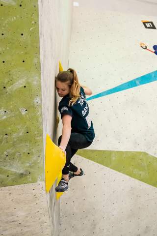 Eine Frau klettert an einer Indoor-Boulderwand und hält sich dabei an großen gelben Griffen fest. Sie trägt enge Sportkleidung und ist konzentriert auf die Kletterroute fokussiert.