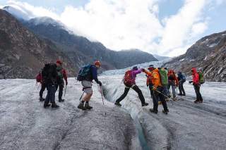 Mehrere mit Wanderausrüstung ausgestattete Personen überqueren einen Gletscher mit sichtbaren Gletscherspalten in gebirgiger Landschaft.