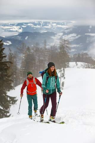 Zwei Personen beim Skitourengehen in verschneiter Berglandschaft mit Nebel und bewaldeten Hügeln im Hintergrund.