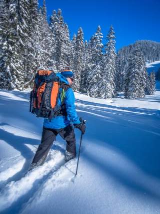 Person mit blauem Winterjacke und großem Rucksack wandert mit Skistöcken durch tiefen Schnee in verschneiter Winterlandschaft mit Tannenbäumen unter blauem Himmel.