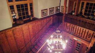 Innenansicht eines Treppenhauses mit dunklem Holzvertäfelung, einer hängenden Kronleuchterlampe und mehreren Fenstern, an den Wänden hängen gerahmte Bilder.