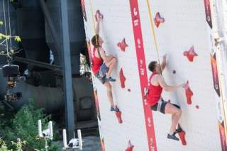 Zwei Kletterer in roten Oberteilen erklimmen eine steile Kletterwand mit roten Griffen.