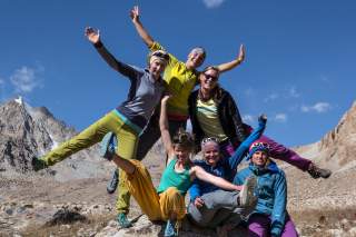 Sechs fröhliche Frauen in bunter Outdoor-Bekleidung posieren spielerisch mit ausgestreckten Armen und Beinen vor einer Berglandschaft unter blauem Himmel.