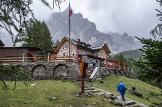 Steinhaus mit rotem Fensterläden und Terrasse, umgeben von Nadelbäumen und Bergen im Hintergrund, Person in blauer Regenjacke auf Treppe im Vordergrund.