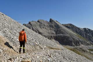 Eine Person mit orangefarbener Jacke und Rucksack wandert auf einem steinigen Hang vor felsigen Bergen unter blauem Himmel.