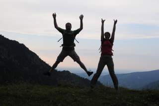 Zwei Personen mit Rucksäcken springen auf einer Bergwiese vor einer bergigen Landschaft und einem bewölkten Himmel in die Luft.