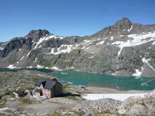 Berglandschaft mit schneebedeckten Gipfeln, einem türkisfarbenen See und einer Berghütte im Vordergrund unter klarem blauem Himmel.
