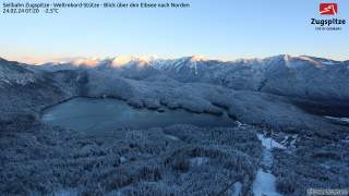 Verschneite Landschaft mit dem zugefrorenen Eibsee im Vordergrund und den von der Morgensonne beleuchteten Bergen der Zugspitze im Hintergrund.