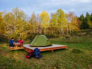 Zwei Personen sitzen an einem Picknicktisch neben einem grünen Zelt auf einer Holzplattform in einer herbstlichen Waldlandschaft, vor ihnen stehen zwei Rucksäcke auf dem Gras.