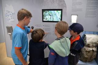Vier Jungen betrachten zusammen einen Bildschirm und eine Wand mit Texten und Bildern in einer Ausstellung.