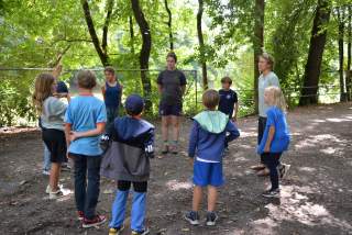 Eine Gruppe von Kindern und zwei Erwachsenen stehen im Kreis im Wald und scheinen an einer Outdoor-Aktivität teilzunehmen. Die Umgebung ist grün und schattig mit vielen Bäumen.