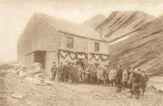 Schwarz-weiß-Foto einer großen Gruppe von Menschen vor einem zweistöckigen Haus mit geschmückter Fassade in einer felsigen Umgebung.