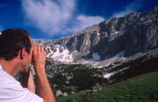 Mann blickt mit Fernglas auf schneebedeckte Berge unter blauem Himmel.