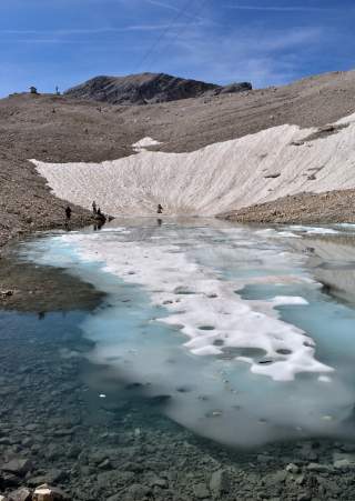 Berglandschaft mit schmelzendem Schnee und Eis auf steinigem Untergrund, dahinter ein felsiger Berg unter blauem Himmel. Menschenn stehen am Rand des Wasserbeckens.