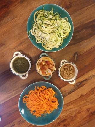 Zwei Teller mit Spiralnudeln aus Zucchini und Süßkartoffel sowie drei kleine Schalen mit Pesto, Tomaten und Zwiebeln auf einem Holztisch.