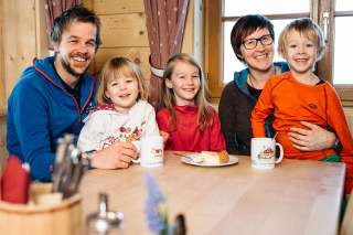 Eine fröhliche Familie mit zwei Erwachsenen und drei Kindern sitzt an einem Holztisch in einer gemütlichen Holzhütte, trinkt aus Tassen und hat ein Stück Kuchen vor sich.