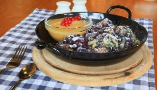 Schwarze Pfanne mit Heidelbeerpfannkuchen und Puderzucker, dazu eine Glasschale mit Apfelmus, garniert mit roten Johannisbeeren, auf Holzuntersetzer und kariertem Tischtuch.