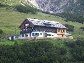 Altes Berghaus mit Holzfassade und bunten Wimpeln auf einer grünen Bergwiese, umgeben von Nadelbäumen und Bergen im Hintergrund.