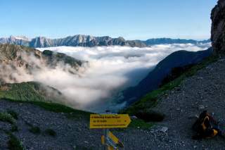Blick von einem Bergpfad auf eine wolkenverhangene Berglandschaft mit schroffen Gipfeln im Hintergrund und einem gelben Wegweiser im Vordergrund.