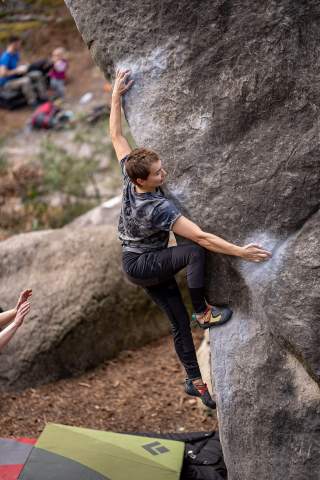 Boulderin Zofia Reych, wie sie aktiv mit großer Anspannung am Felsen klettert. Die Griffpunkte am Fels sind von weißen Chalk gezeichnet. Unterhalb liegt ein grünes Crashpad, was einen eventuellen Fall abfedern soll.