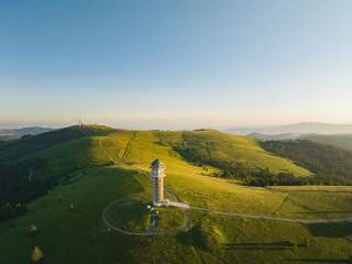 Aussichtsturm auf dem Feldberg im Schwarzwald, umgeben von Wiesen und Wäldern, im Hintergrund weitere Hügel und Berge unter klarem Himmel.