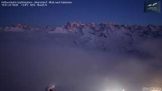 Schneebedeckte Berggipfel bei Dämmerung über einer dichten Wolkenschicht, unterhalb sind Lichter einer Siedlung sichtbar. Die Temperatur beträgt -13,8°C, der Himmel ist klar.