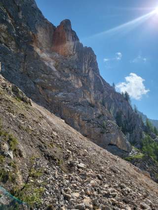 Steiler Felsabhang mit Geröll unter klarem, sonnigem Himmel und wenigen Wolken. Im Hintergrund sind vereinzelt kleine Bäume an der Felswand zu sehen.