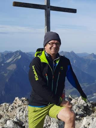 Stefan Wagegg sitzt vor einem Gipfelkreuz im Hintergrund Berggipfel.