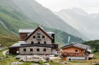 Steinhütte und Holzhütte inmitten grüner Berglandschaft, im Hintergrund nebelverhangene Berge.