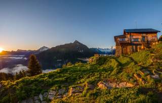 Berglandschaft bei Sonnenuntergang mit einer Hütte auf einer grünen Wiese im Vordergrund und schneebedeckten Gipfeln im Hintergrund.