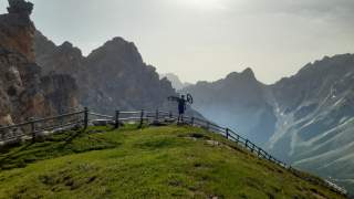 Person mit Mountainbike auf den Schultern steht auf einer mit Gras bewachsenen Kuppe mit Blick auf die umliegenden Felsgipfel