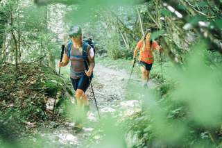 Zwei Frauen wandern mit Trekkingstöcken auf einem schmalen Waldweg, umgeben von dichtem grünem Laub.