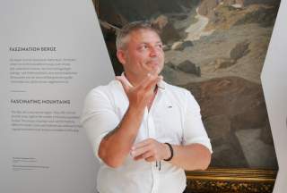Ein Mann in weißem Hemd steht vor einer Ausstellungstafel und einem Gemälde, er gestikuliert mit den Händen und blickt nach rechts.